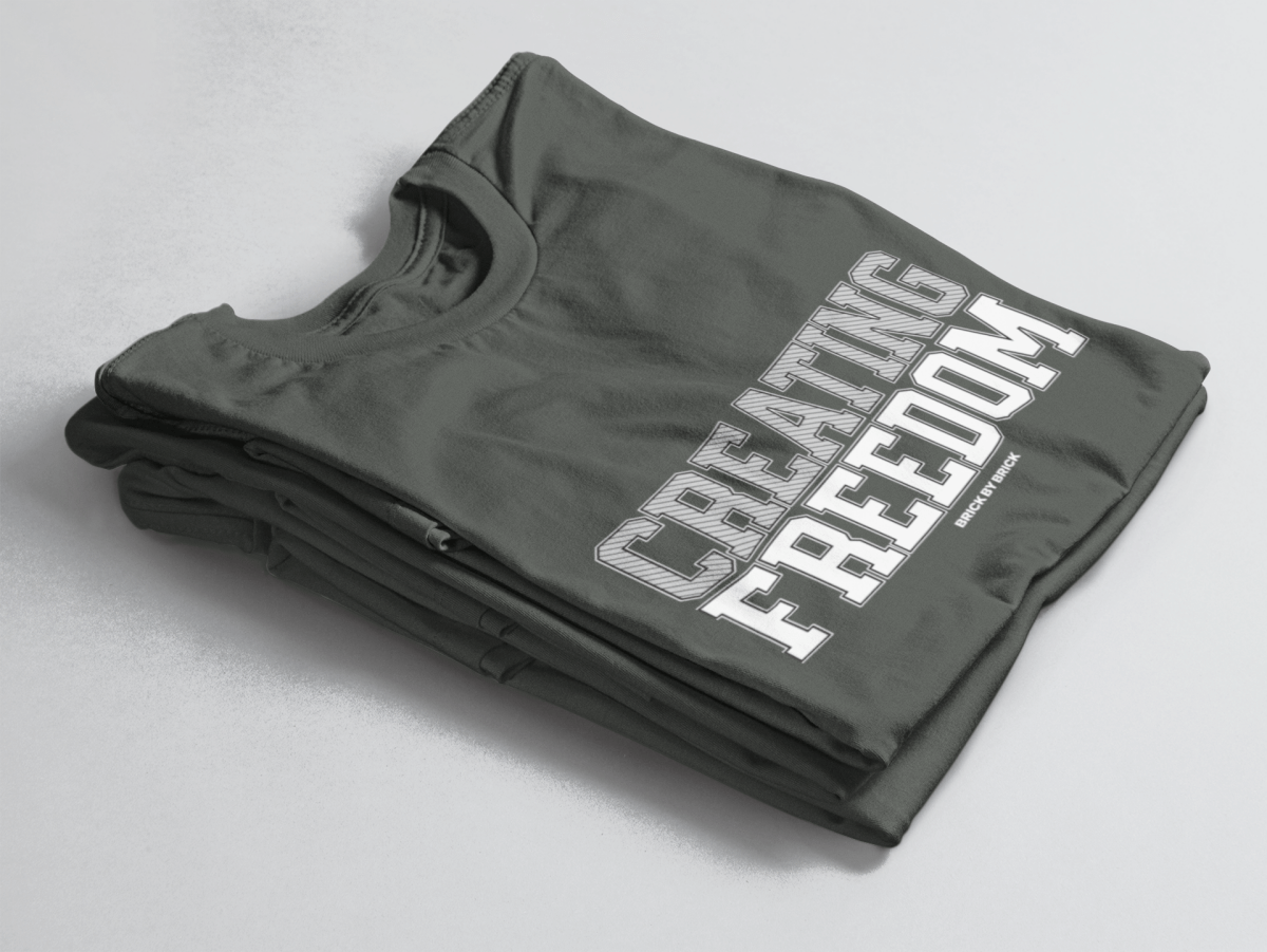 Charcoal Green folded tee with bold gray and white text “Creating Freedom” and small “Brick by Brick” centered below