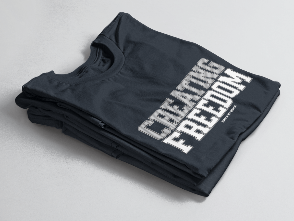 Navy folded tee with bold gray and white text “Creating Freedom” and small “Brick by Brick” centered below