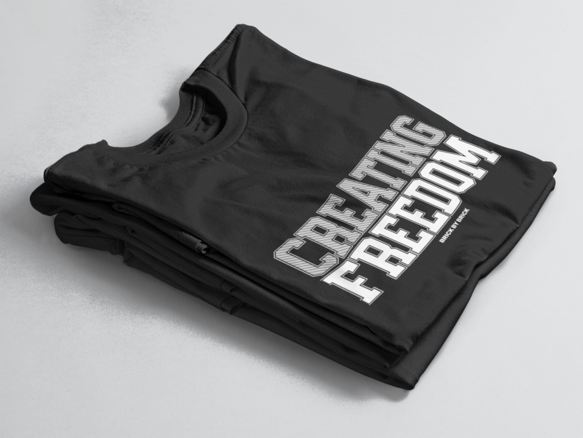 Black folded tee with bold gray and white text “Creating Freedom” and small “Brick by Brick” centered below.