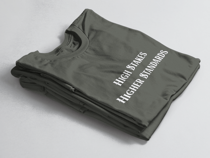 Charcoal green folded tee with white gothic-style text reading “High Stakes Higher Standards” centered on the front.