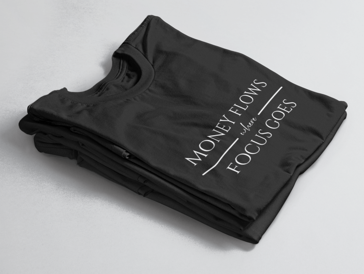 Black folded tee with white text “Money Flows Where Focus Goes” in elegant serif font, with horizontal lines next to “where"