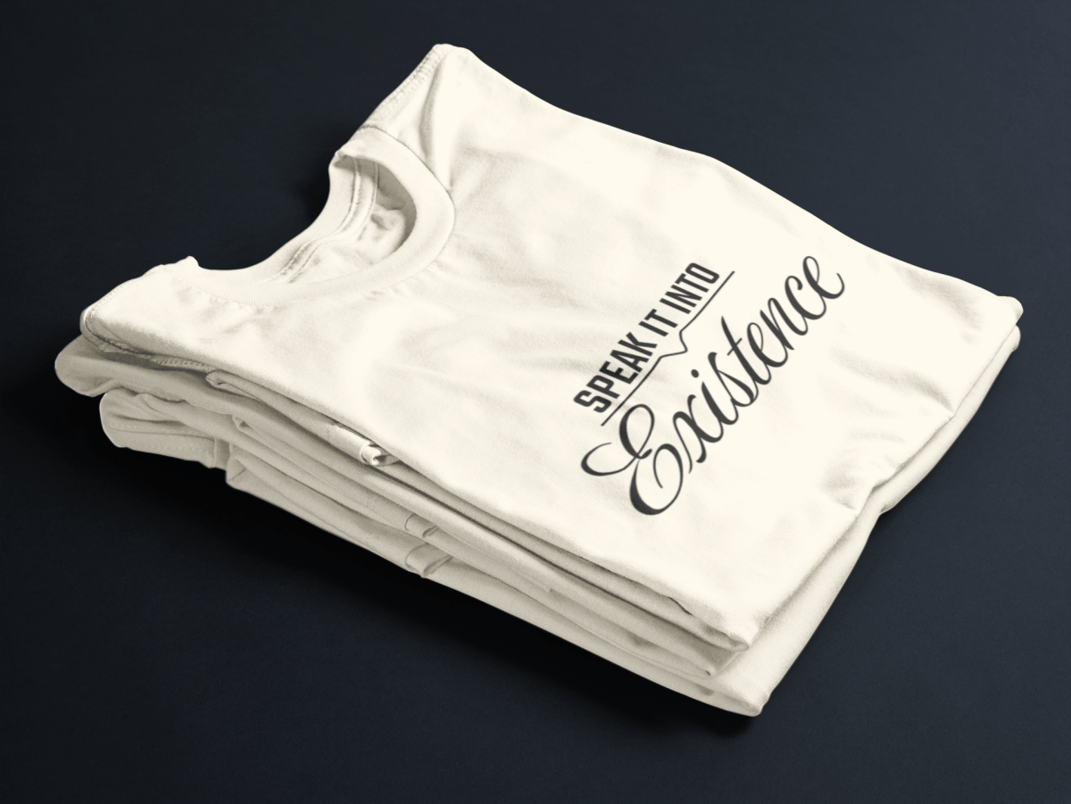 Beige folded tee with black text “Speak It Into” in bold caps above cursive “Existence” centered on the front.
