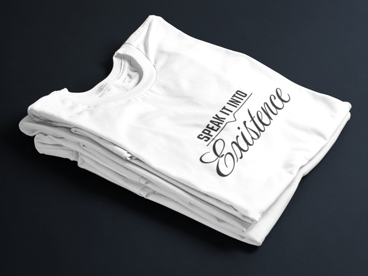 White folded tee with black text “Speak It Into” in bold caps above cursive “Existence” centered on the front.