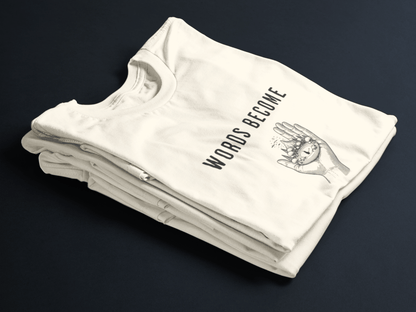 Beige folded tee with black text “Words Become Worlds” and a hand holding a tiny illustrated world in the center