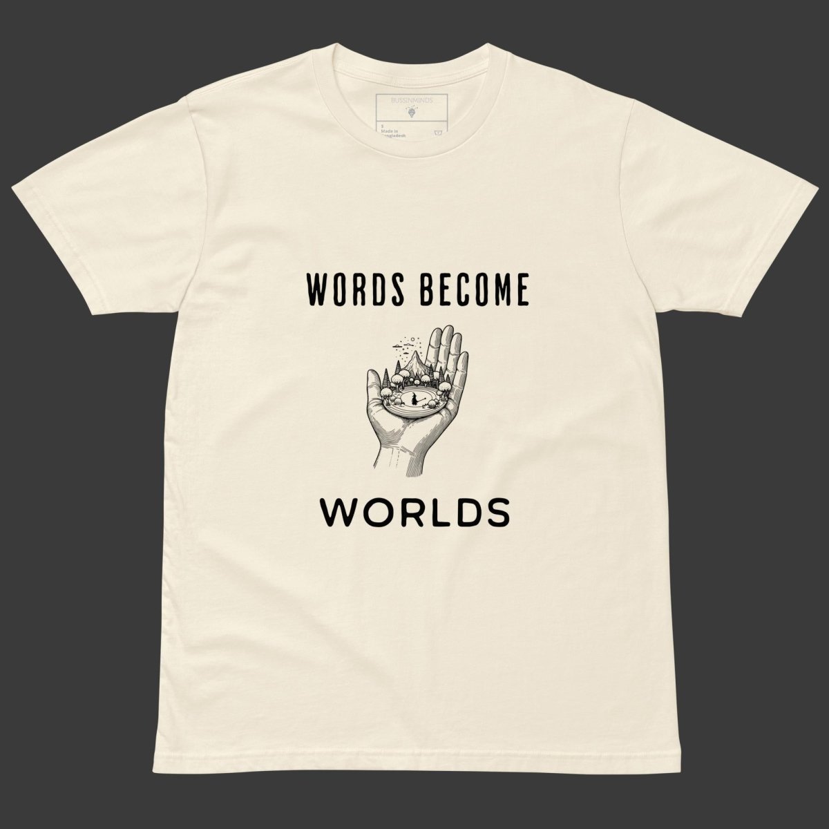 Beige tee with black text “Words Become Worlds” and a hand holding a tiny illustrated world in the center