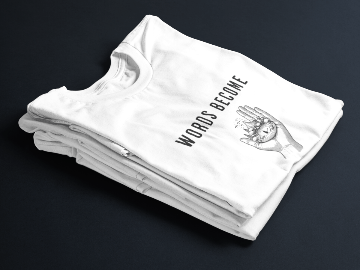 White folded tee with black text “Words Become Worlds” and a hand holding a tiny illustrated world in the center