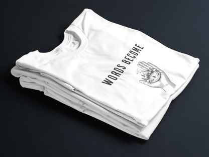 White folded tee with black text “Words Become Worlds” and a hand holding a tiny illustrated world in the center