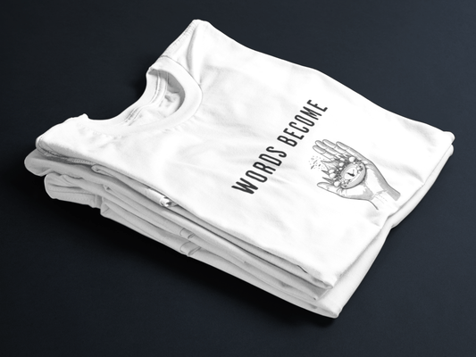 White folded tee with black text “Words Become Worlds” and a hand holding a tiny illustrated world in the center